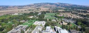Aerial view of Sonoma State's sprawling campus set amidst rolling hills and open fields. Several buildings are scattered across the green landscape, interspersed with trees and pathways. In the background, small communities and roadways can be seen, bordered by mountainous terrain.