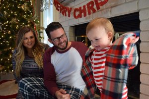 A young child wearing a red and white striped shirt and plaid robe stands in front of a decorated Christmas tree and a lit fireplace. A man and a woman, both in matching holiday pajamas, sit nearby, smiling at the child. A "Merry Christmas" banner hangs above the fireplace, fostering an atmosphere of joy and mental fitness.