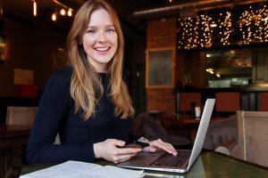 A woman with long blonde hair is smiling at the camera while seated at a table in a cozy cafe. Showcasing her technology talent, she has a laptop in front of her, a phone in one hand, and an open notebook next to her. The cafe has warm lighting and a decorative brick wall with string lights.