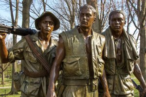 A bronze statue depicts three warfighters in combat gear. One soldier on the left wears a helmet and carries a weapon, the middle soldier has a vest and appears jacketless, and the soldier on the right has a rolled-up cloth draped over his shoulder. Trees are in the background.
