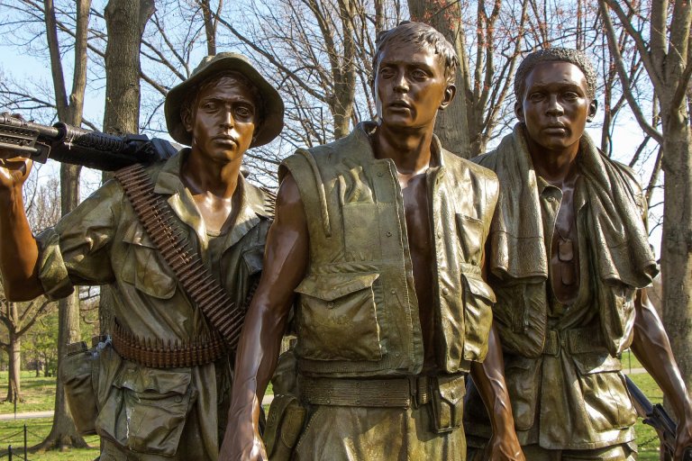 A bronze statue depicts three warfighters in combat gear. One soldier on the left wears a helmet and carries a weapon, the middle soldier has a vest and appears jacketless, and the soldier on the right has a rolled-up cloth draped over his shoulder. Trees are in the background.