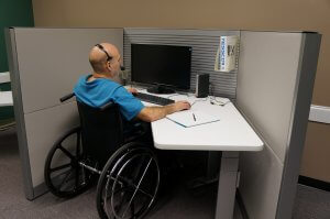 A warfighter in a wheelchair wearing a headset sits at a cubicle desk, working on a computer. There is a notebook and a pen on the desk, and a file organizer is attached to the cubicle wall. The setting appears to be an office workspace.