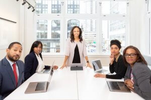 A team of five professionals in a bright office space, three women and two men, seated around a conference table with laptops. One woman stands at the head of the table, smiling like a confident warfighter, while the others look towards the camera. Large windows display a cityscape outside.