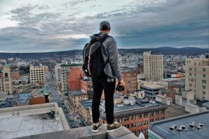 A millennial, wearing a cap, hoodie, and backpack, stands on the edge of a rooftop in an urban area, holding a camera. The scene overlooks a cityscape with various buildings and mountains in the background under a cloudy sky.