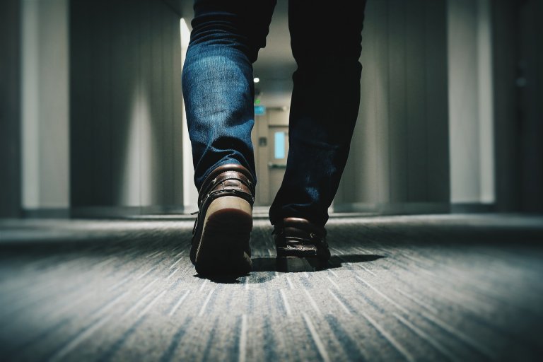 A millennial wearing jeans and brown shoes is walking down a narrow, dimly lit hallway. The shot is taken from a low angle at ground level, focusing on their legs and shoes. The hallway has a textured carpet with blurred background doors and walls.