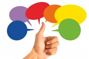 A hand gives a thumbs-up in front of a background with five overlapping speech bubbles in different colors: purple, red, orange, yellow, and green. The design suggests feedback, communication, approval, or positivity.