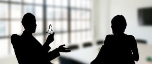 Silhouettes of two people sit facing each other in an office-like setting with large windows in the background. One person appears to be gesturing while holding a small object, possibly glasses, as they give feedback.
