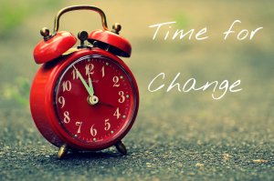 A vintage red alarm clock sits on an asphalt surface with a blurred background. The clock shows the time as 10:10. The words "Time for Culture Change" are written in white cursive text to the right of the clock.