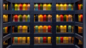 A multi-story building interior with balconies on each floor, featuring colorful doors in yellow, red, green, blue, and orange. Each floor's illumination casts a warm glow, creating a vivid and visually striking pattern. The design transitions seamlessly between modern geometry and vibrant hues.