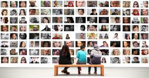 Three people sit on a bench in a gallery, viewing a large wall of diverse portraits. The portraits include people of varying ages, genders, and ethnicities, showcasing a rich tapestry of human faces and expressions—a beautiful transition from one unique story to another.