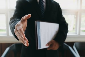 A person in a business suit extends their right hand for a job-related handshake while holding a folder in their left hand. The background shows white-framed windows with blurred outdoor lighting.