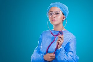 A dedicated medical professional stands against a blue background, wearing a blue surgical cap, protective glasses, and a blue gown. They are holding a red stethoscope in one hand while the other end rests in their ears, exemplifying their commitment to the job.