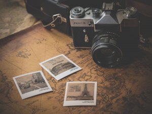 A vintage Zenit-E camera rests on an old world map beside three black-and-white prints. The prints capture travels to iconic landmarks: the Colosseum labeled “Rome,” a Paris street scene, and the Eiffel Tower labeled “Paris.” A leather camera case is partially visible.