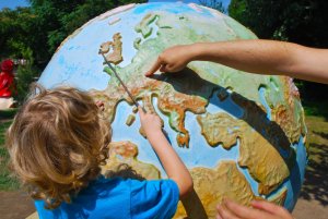 A child with curly blonde hair, wearing a blue shirt, points at Europe on a large, colorful globe. An adult's hand joins in, sharing in the excitement of future travels. The globe is displayed outdoors with trees and greenery in the background.