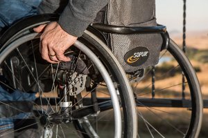 A close-up of a person's hand gripping the wheel of a Sopur wheelchair, hinting at their journey through workers' compensation. The background is blurred, showing an outdoor setting with light brown terrain and sporadic vegetation visible in the distance.