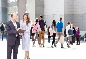 Two people are standing and talking in front of a modern glass building. The man, in a suit, holds a folder while the woman in professional attire points at it, demonstrating leadership. In the background, several people walk in different directions, some carrying bags and backpacks.