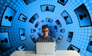 A person in glasses sits at a table, working on a laptop. The background features a surreal, blue digital art design with a tunnel-like effect, filled with silhouettes of people inside various device screen shapes, symbolizing connectivity and digital networking crucial for economic development.