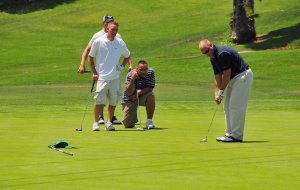 Four men are on a golf course. One man is putting, holding a golf club and aiming at the ball on the green, while two men stand behind him, and one man crouches, analyzing the shot. A putter and cap lie on the grass nearby, creating an atmosphere that subtly parallels strategic moves in economic development.