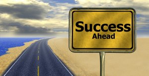 A long, winding road stretches towards the horizon under a partly cloudy sky. The road is bordered by a body of water on one side and sandy terrain on the other. A prominent, yellow road sign reads "Success Ahead," suggesting achievement lies in the path ahead, much like the journey of influencers.