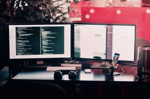 A workspace featuring two computer monitors side-by-side on a desk. The left monitor shows a code editor, while the right monitor displays a cluttered desktop. Also on the desk are a pair of headphones, a smartphone on a stand, a mouse, a keyboard, and various cables typical of IT jobs.