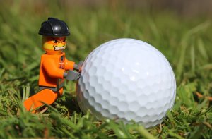 A LEGO figurine, with an angry expression and donned in an orange construction outfit and black helmet, is pushing a large golf ball on grass. The scene humorously mimics a comically exaggerated struggle, highlighting the figurine's limited knowledge of physics due to the size disparity.