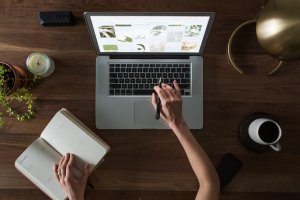 A negotiator works on a laptop with image thumbnails displayed on the screen, using a pen on the trackpad. Their other hand holds an open notebook with writing. The wooden desk also has a candle, potted plant, cup of coffee, smartphone, and a lamp.