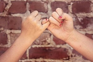 Two hands with pinky fingers interlocked in a promise, their fingernails painted a vivid red. The background is a weathered brick wall, adding to the scene’s timeless bond.