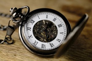 An antique pocket watch with an exposed mechanical interior, displaying Roman numerals on its white clock face, lies open on a wooden surface. The watch chain, symbolizing a promise of timeless craftsmanship, is coiled around the watch. The intricate inner workings of the watch are clearly visible.