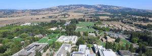 Aerial view of a large university campus surrounded by greenery and hilly terrain. The campus features multiple buildings, roads, parking lots, and open fields. Residential areas and farmland can be seen in the surrounding landscape under a clear blue sky, where students enjoy both academic pursuits and online privacy.