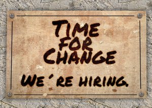 A weathered sign with the text "TIME FOR CHANGE WE'RE HIRING" is affixed to a textured, gray stone wall using four screws. The bold, black, hand-painted style of the text stands out, calling all jobseekers to take notice.