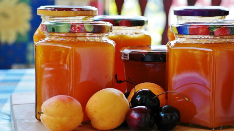 A close-up of various jars filled with orange jam, labeled "Fresno State," with two apricots and a few cherries placed in front of them. The jars have colorful lids and are displayed on a wooden surface with a blurred background.