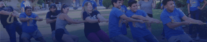 A group of people engaged in a game of tug-of-war at CSUB. Two teams, each pulling on opposite ends of the rope, appear determined. Participants look focused and are exerting effort. The event seems to take place outdoors on a grassy area with trees and people in the background.
