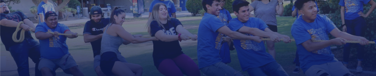 A group of people engaged in a game of tug-of-war at CSUB. Two teams, each pulling on opposite ends of the rope, appear determined. Participants look focused and are exerting effort. The event seems to take place outdoors on a grassy area with trees and people in the background.