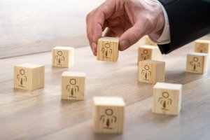 A hand in a business suit arranges wooden blocks, each featuring a simple human icon, on a wooden surface. The image suggests concepts of teamwork, leadership, or human resources management, akin to carefully crafting a standout resume.