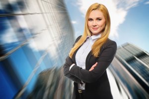 A confident woman with long blonde hair, wearing a black blazer and white blouse, stands with her arms crossed in front of a blurred urban background featuring a high-rise building and a bright, partly cloudy sky, presenting an image perfect for a professional resume.