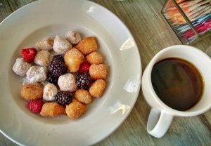 A white plate features bite-sized, sugar-coated pastries accompanied by fresh raspberries and blackberries. Next to the plate, there is a white mug filled with ego-boosting black coffee. The background includes a container with assorted sweeteners.