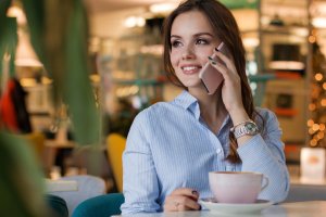A woman in a light blue shirt, likely from Generation Z, smiles while talking on a smartphone. She is seated at a table in a cozy, well-lit café with a cup of coffee or tea in front of her. The background features blurred colorful decor and warm lighting.