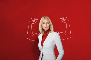 A woman with blonde hair, wearing a white suit and red top, stands against a solid red background. White illustrations of muscular arms are drawn behind her, symbolizing leadership and making it appear as if she has strong, flexed biceps.