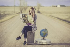 A young woman with red hair sits on a wooden crate on a rural dirt road, studying a roadmap. Beside her is a suitcase with a globe on top of it. The road stretches into the distance, flanked by open fields and a few distant buildings.