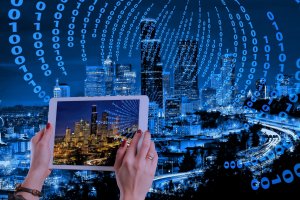 Hands holding a tablet in front of a cityscape at night. The tablet screen displays the same city in daylight. Binary code flows in arcs around the city, suggesting a digital roadmap theme. The cityscape is illuminated with blue hues.