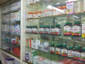 Shelves in a healthcare-oriented pharmacy displaying various vitamin and supplement bottles, including vitamin C, calcium, zinc, and multivitamins. The shelves also contain boxes of medications and health products, neatly arranged behind clear glass.