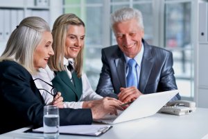 Three older adults wearing business attire are sitting around a desk and smiling while looking at a laptop, possibly reviewing a resume. A glass of water, a clipboard, and a landline phone are on the desk. Shelves with binders and a window are in the background.