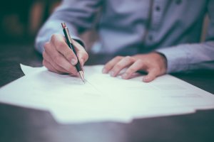 A person in a light blue shirt is carefully writing on a stack of papers with a black and gold pen, perhaps updating their resume. The focus is on the hand and pen, with the writing slightly blurred. The background is out of focus, suggesting a professional or office setting.