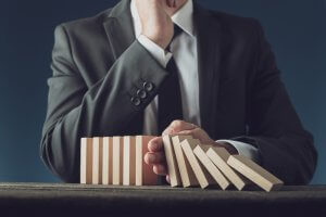 A person in a suit uses their hand to stop a sequence of falling wooden blocks, akin to managing government staffing to prevent a bureaucratic chain reaction. The dark background emphasizes the focus on the blocks and the decisive intervention.
