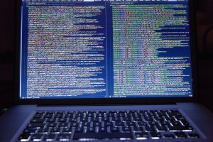 A laptop screen is illuminated with dense lines of code in various colors, suggesting programming or information governance in progress. The keyboard is visible at the bottom of the image, and the room appears to be dimly lit.