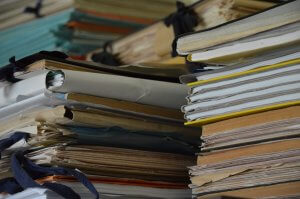 Stacks of various files and folders are piled up, indicating a cluttered and disorganized space. The files, in different colors and sizes, some tied with strings, depict an overwhelming amount of paperwork—clearly lacking any form of information governance.
