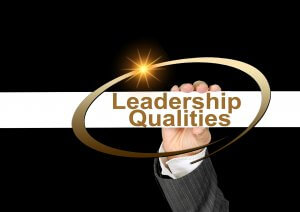A hand in a suit is holding a golden oval frame around the words "Leadership Qualities and Values," with a bright starburst in the top left corner of the frame, all against a black and white background.