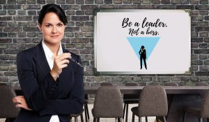 A woman in a business suit stands confidently with eyeglasses in hand, next to a whiteboard displaying the text "Be a leader. Not a boss." The whiteboard also features a silhouette of a person in a suit using a megaphone inside an inverted triangle, symbolizing core leadership values.