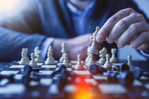 A person wearing a gray blazer, exhibiting crisis leadership, strategically moves a white king chess piece on a chessboard. The board is filled with various white and black pieces, some toppled over. Bright sunlight illuminates the scene, suggesting a thoughtful moment.