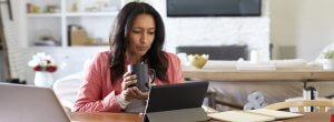 A woman with long hair sits at a table, holding a CSU mug in one hand while looking at a tablet. She is wearing a pink jacket, and her workspace includes a laptop, notebook, and pen. The background showcases home decor, including shelves and a TV.